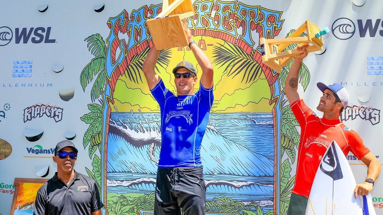 Three surfers posing on a competition podium with raised arms in front of a colorful event banner
