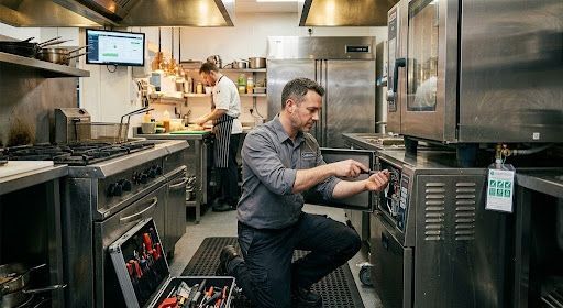 Chef working in a stainless-steel restaurant kitchen, adjusting a machine beside a stovetop.