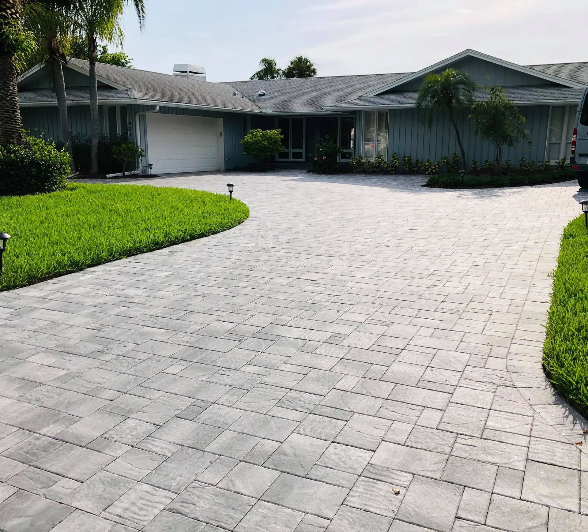 Wide brick paved driveway leading to a single-story gray house with green lawn and palm trees