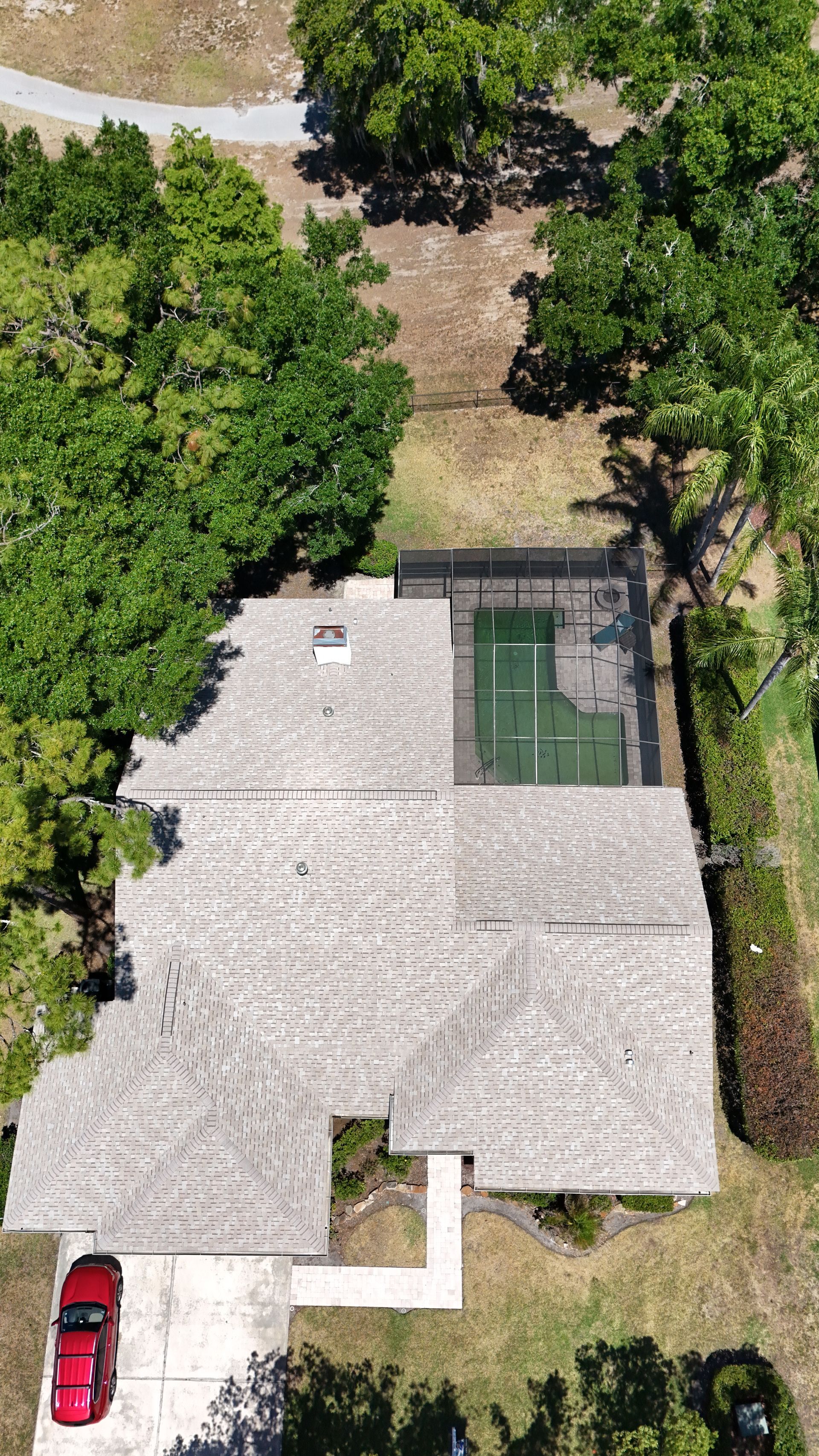 Aerial view of a house with a light roof, backyard pool, driveway, and surrounding trees.