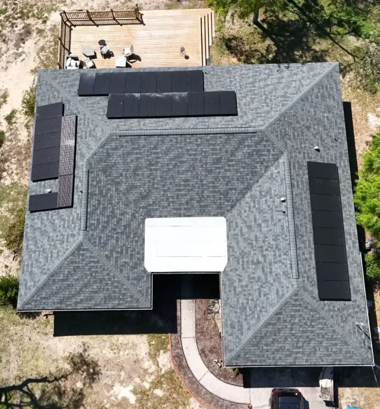 Aerial view of a gray-shingled house with a white awning, black roof panels, and a patio in the yard
