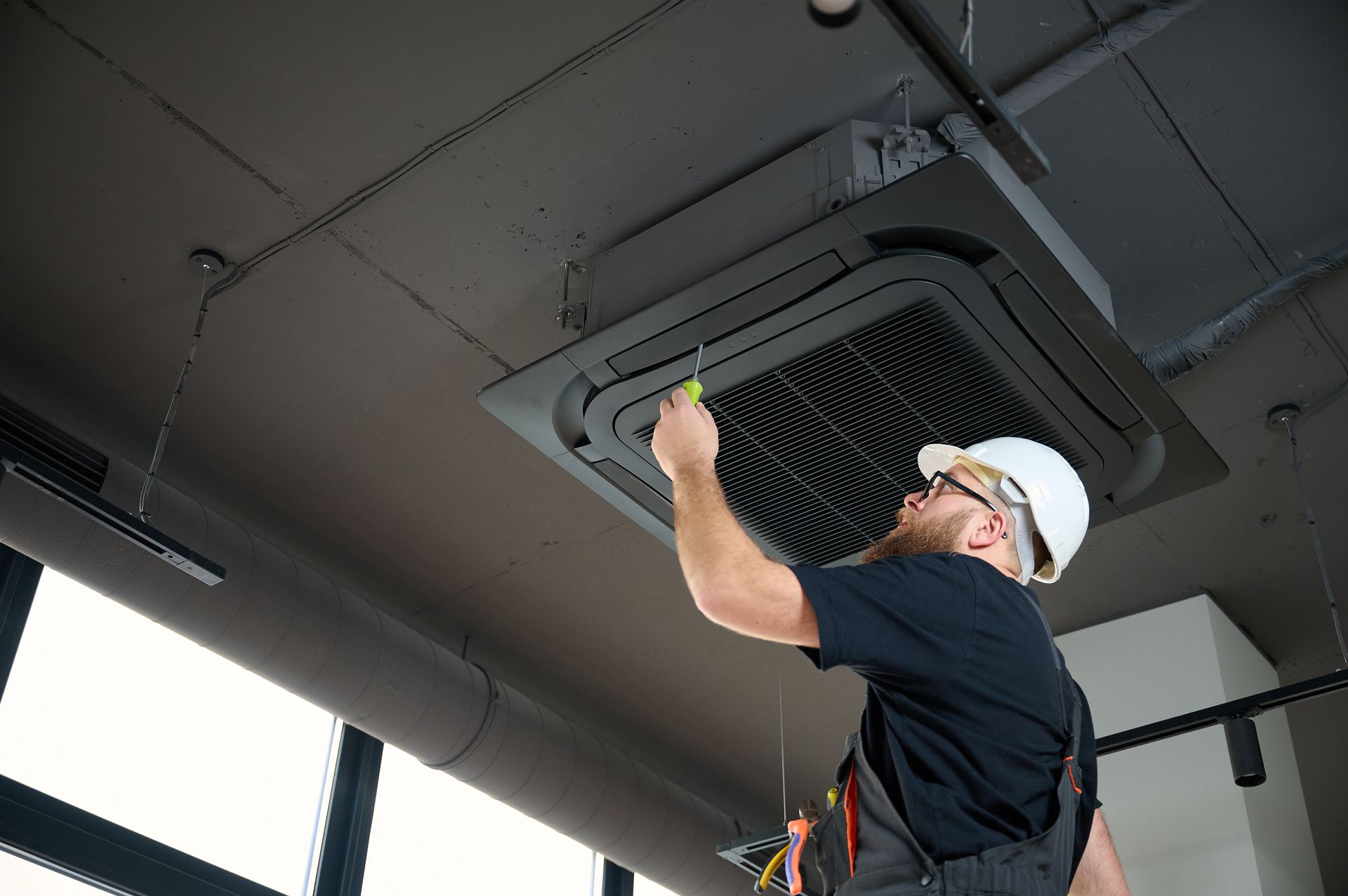 HVAC technician in a hard hat, working on a ceiling-mounted air conditioning unit with a screwdriver.