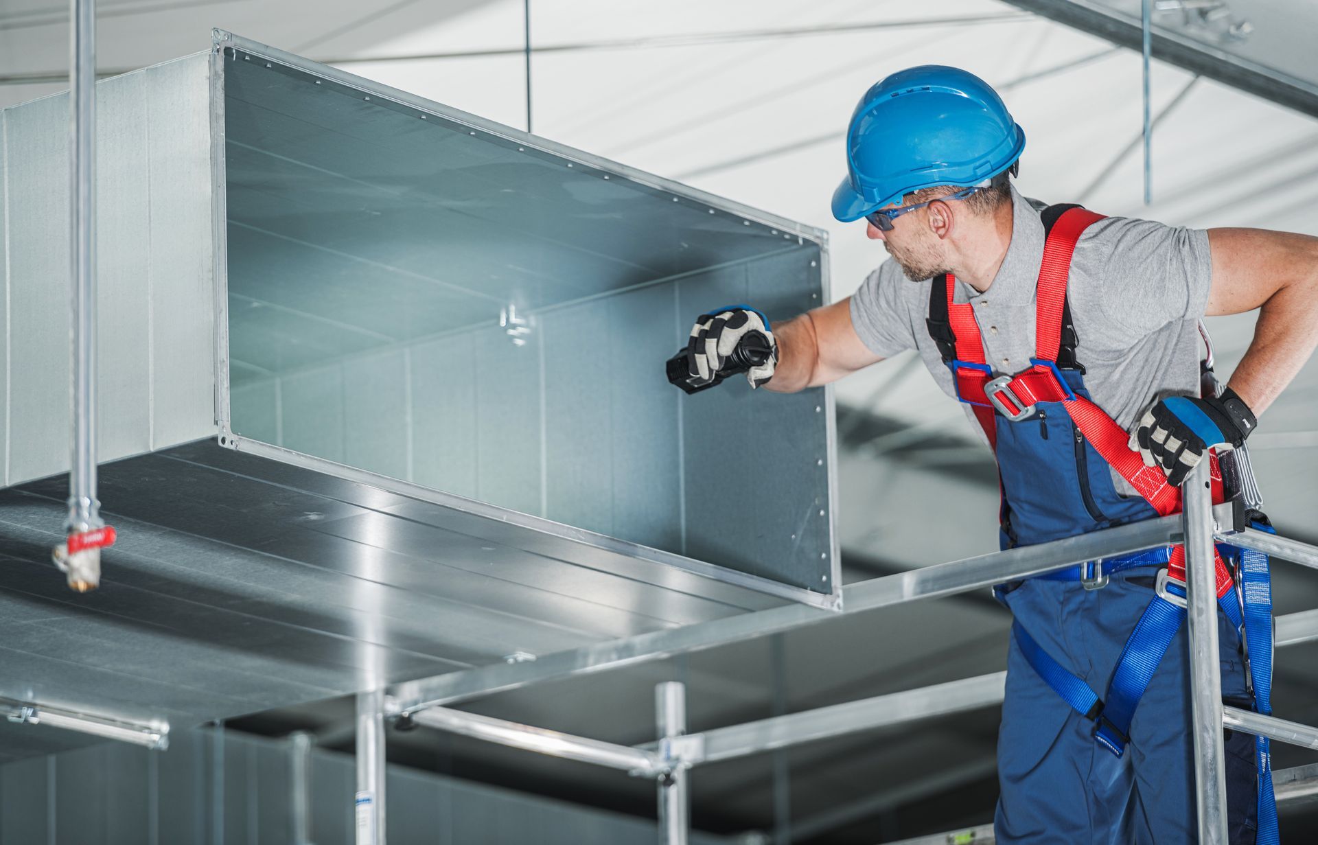 HVAC worker in blue overalls and hard hat on scaffolding, inspecting ventilation duct.