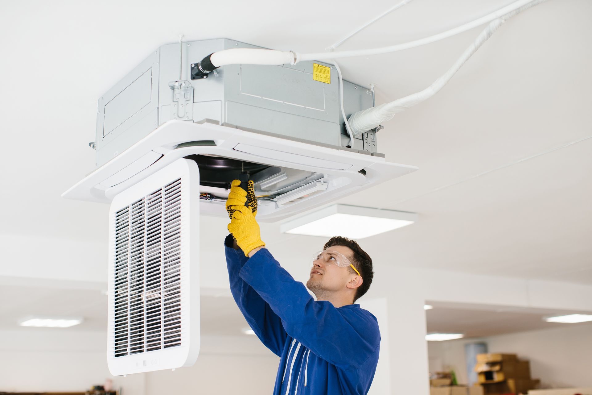 Technician in blue coveralls fixing a ceiling-mounted HVAC unit with yellow gloves, in a white room.