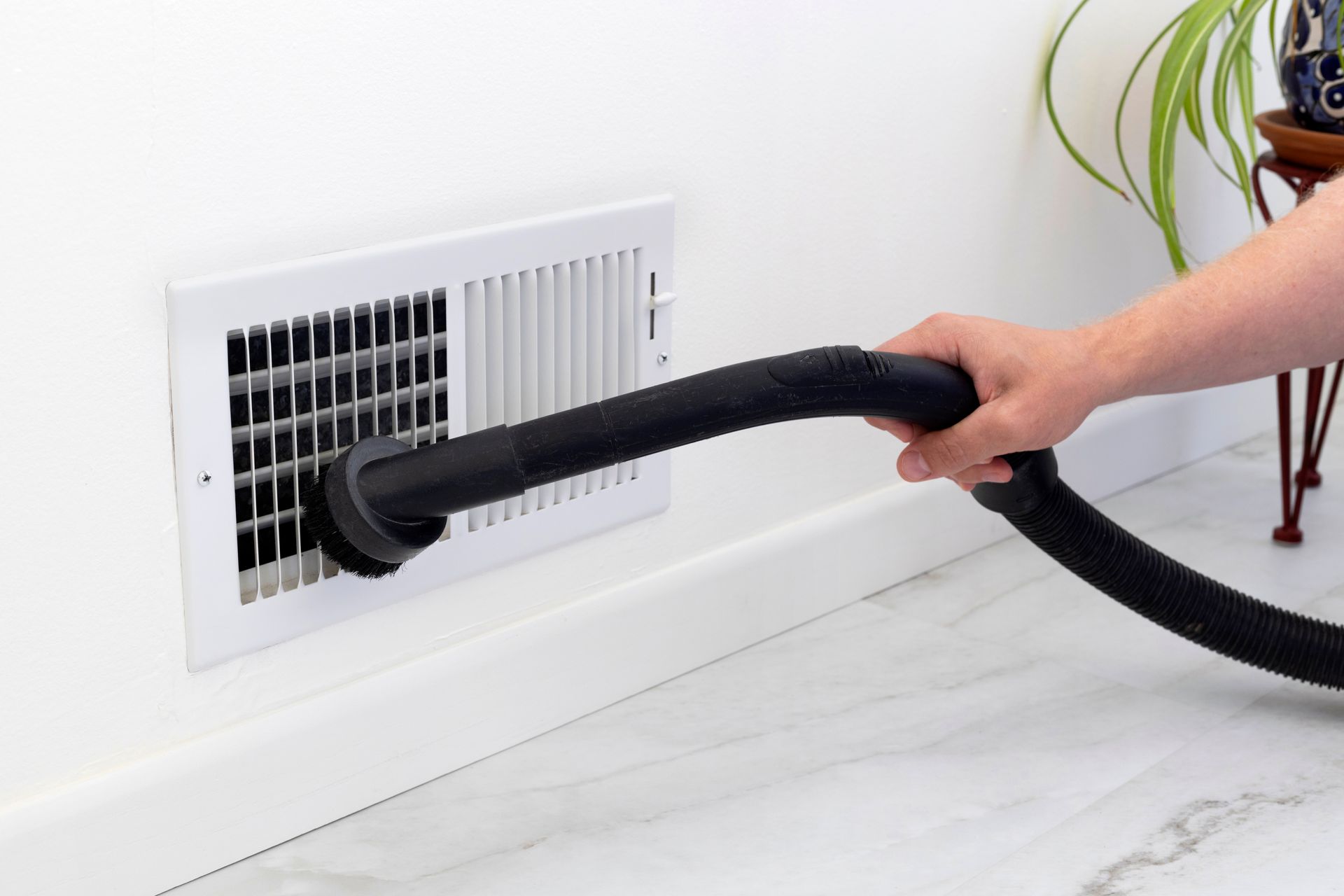 A person vacuums a vent on a white wall, using a black hose.