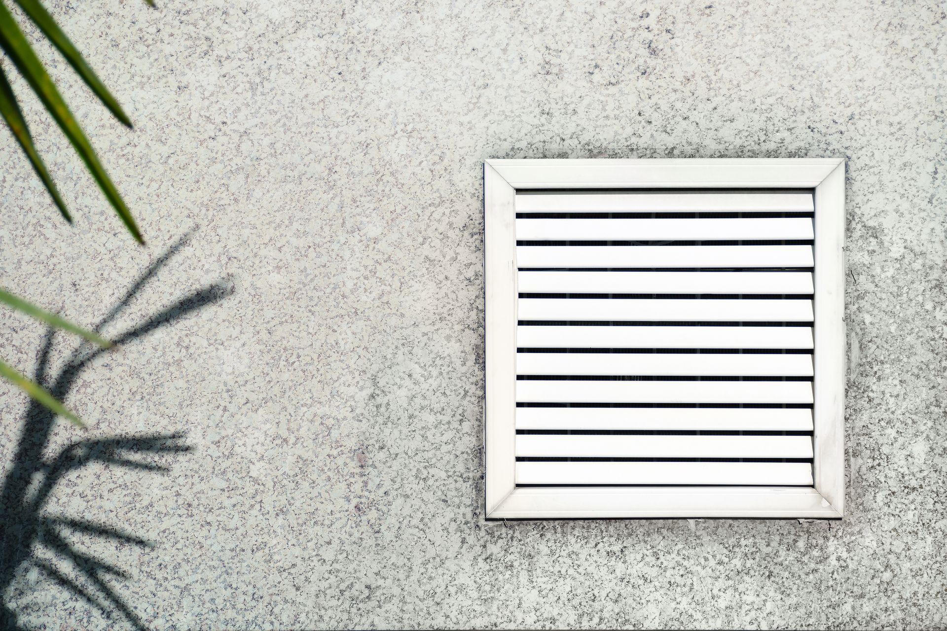 White vent with horizontal slats on a textured gray wall; plant shadow in the left corner.