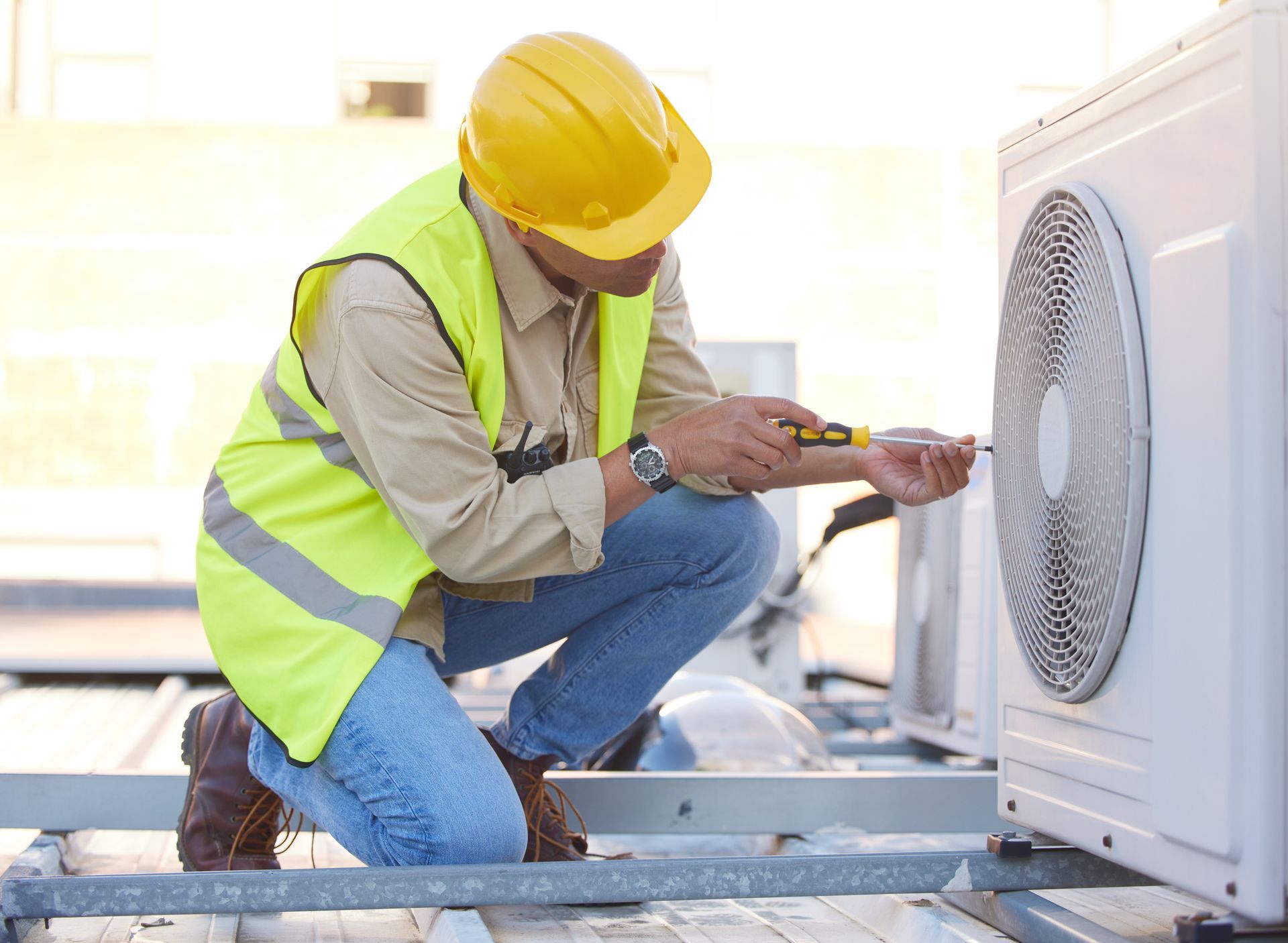 HVAC technician in yellow hard hat and vest, kneeling and working on outdoor air conditioning unit.