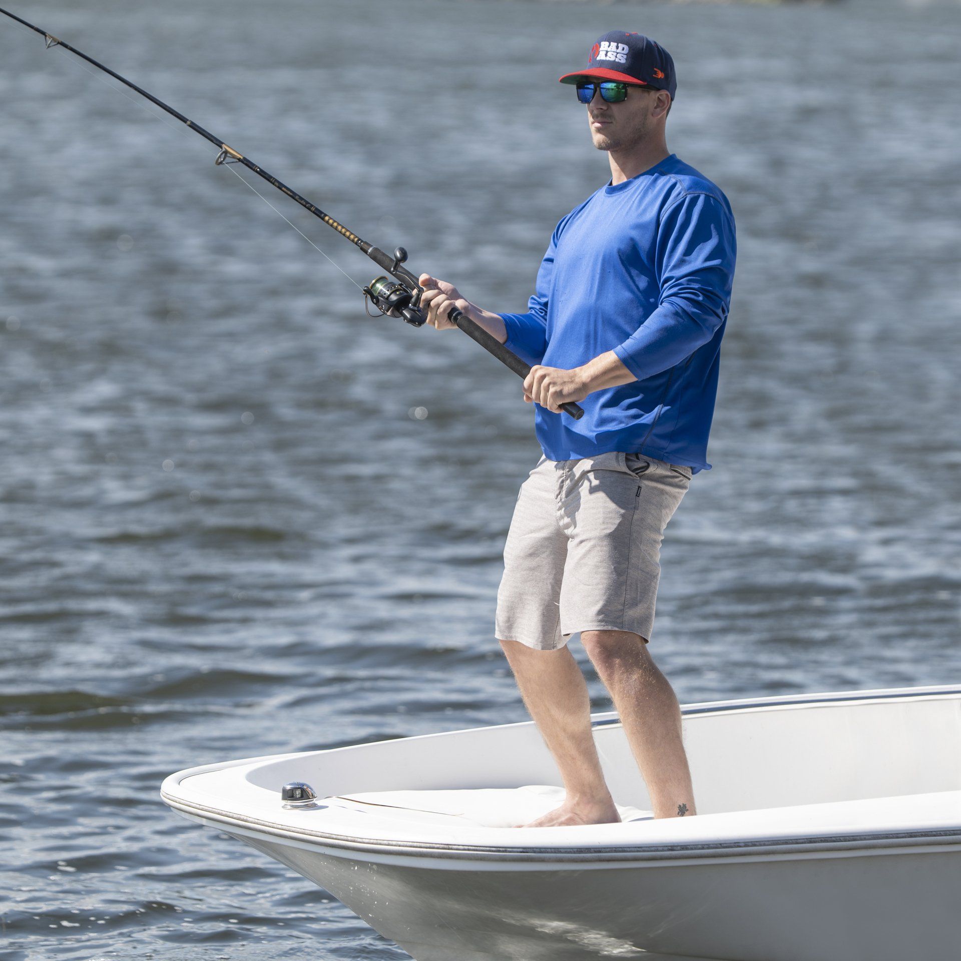 Man fishing from a boat, wearing a blue shirt, khaki shorts, and a baseball cap on a sunny day.