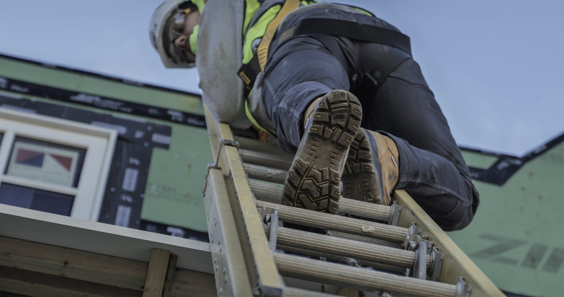 Construction worker wearing safety gear climbing a ladder to the roof of a building.