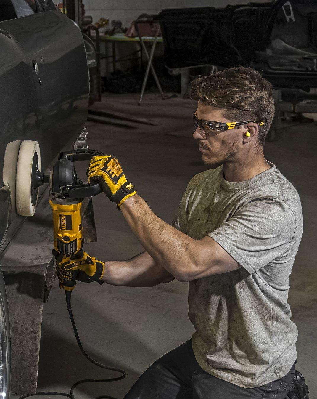 Man kneels, polishing a car with a power buffer. He wears safety glasses, gloves, and ear plugs in a garage.