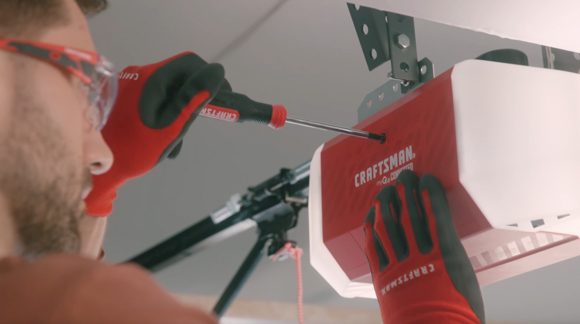 Man in red gloves, safety glasses, installing garage door opener with a screwdriver.