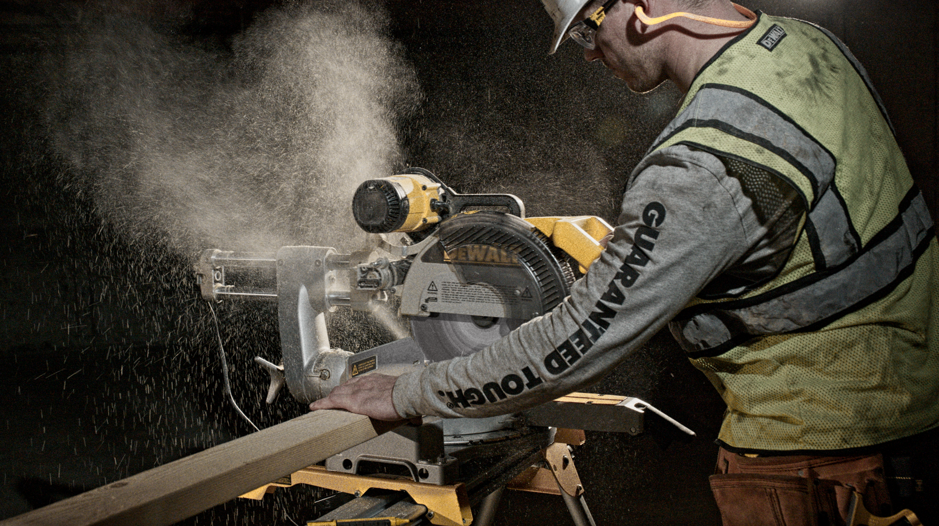 Construction worker using a miter saw, creating dust. Worker is wearing safety gear and is inside a dark setting.