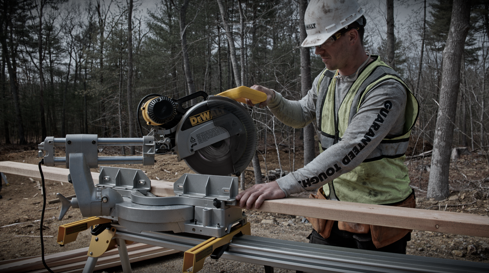 Man in safety gear using a miter saw outdoors to cut wood.