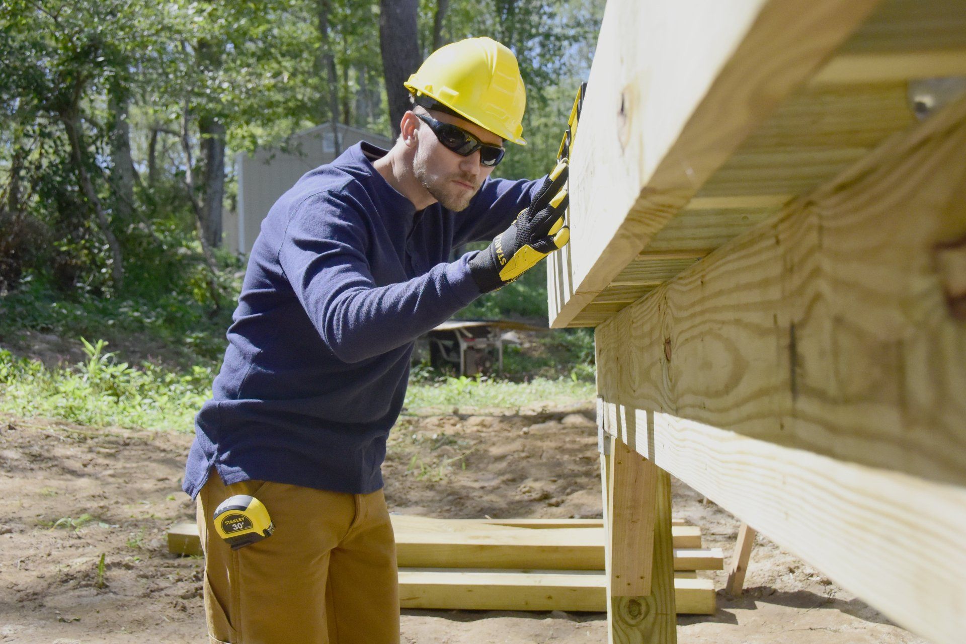 Construction worker in a yellow hard hat, sunglasses, and work gloves measures wooden deck with a level.