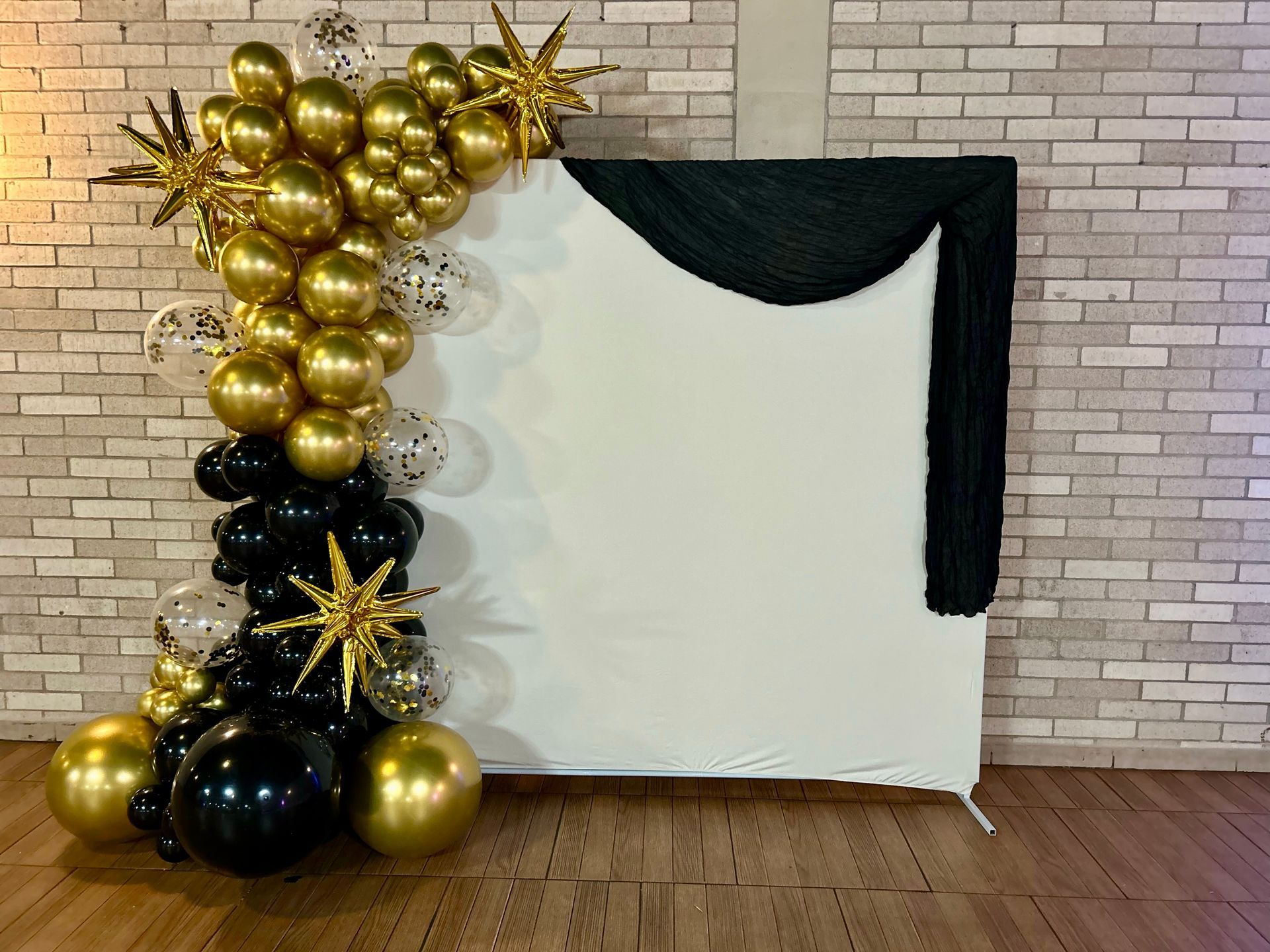 Gold and black balloon arch with a white backdrop and black fabric. Brick wall and wood floor.