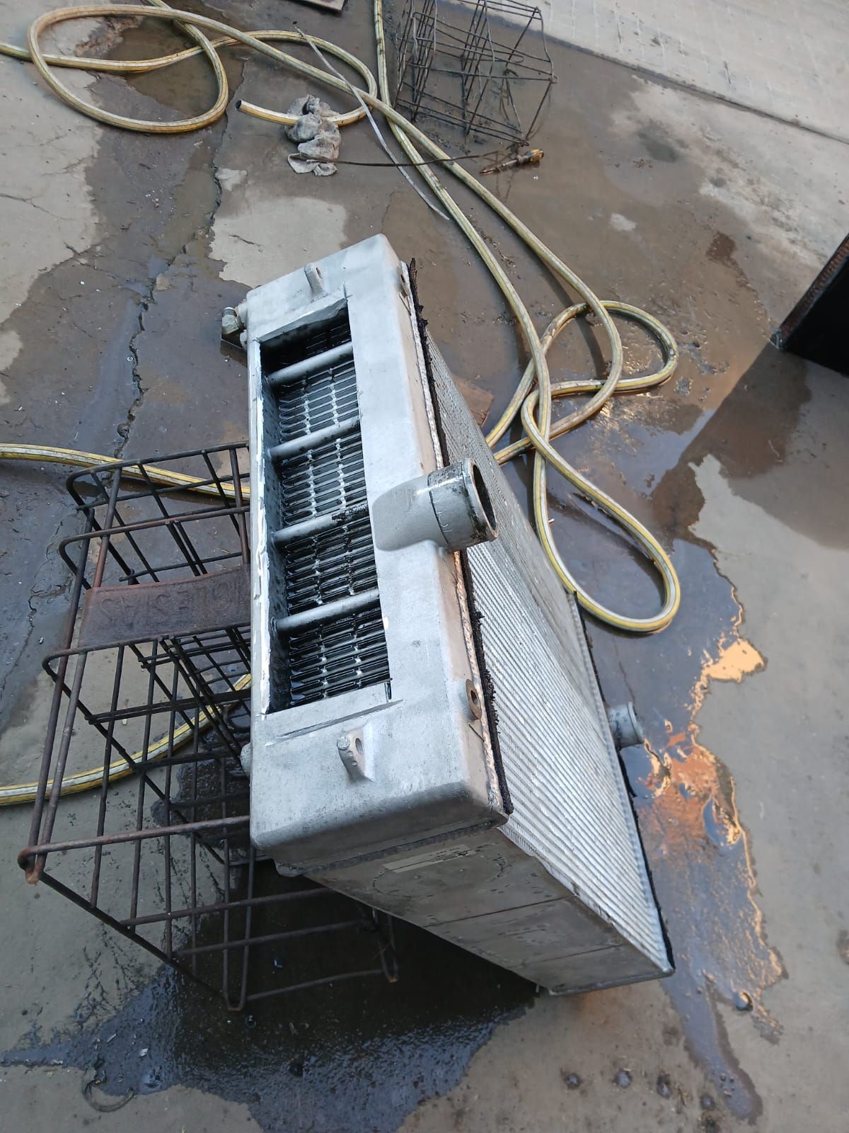 Una unidad de aire acondicionado averiada yace sobre concreto húmedo junto a un carrito de metal y una manguera enrollada.
