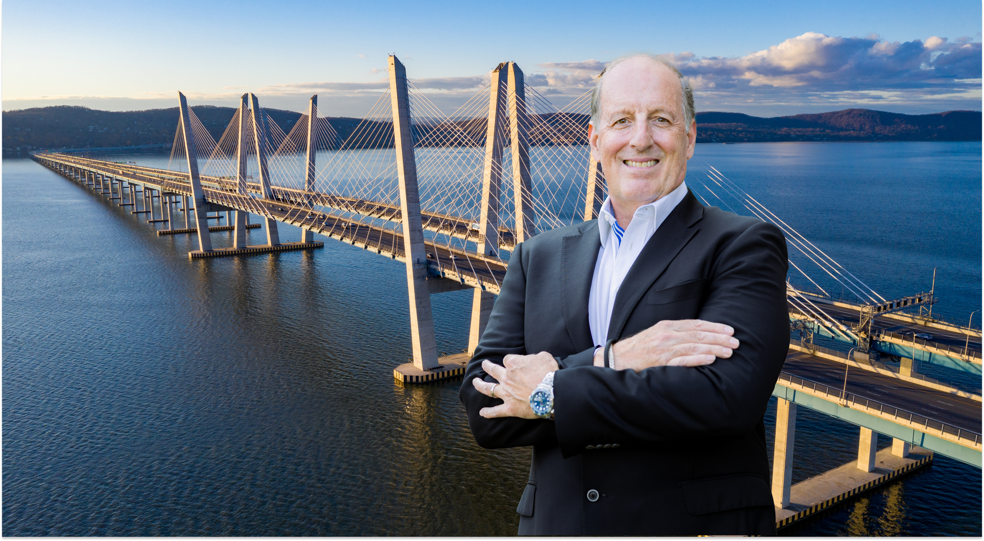 Man in suit stands with arms crossed in front of the Governor Mario M. Cuomo Bridge at sunset.