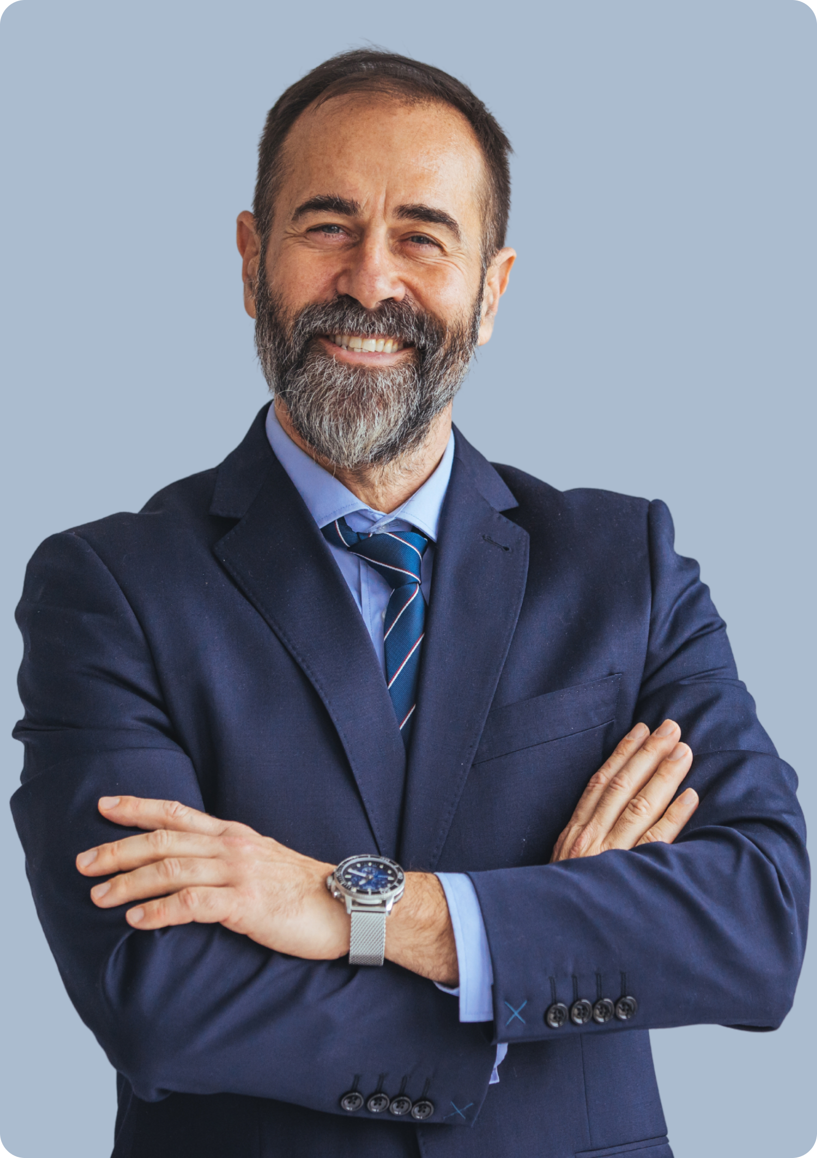 Man in navy suit with crossed arms, smiling. Grey beard, watch. Pale blue background.