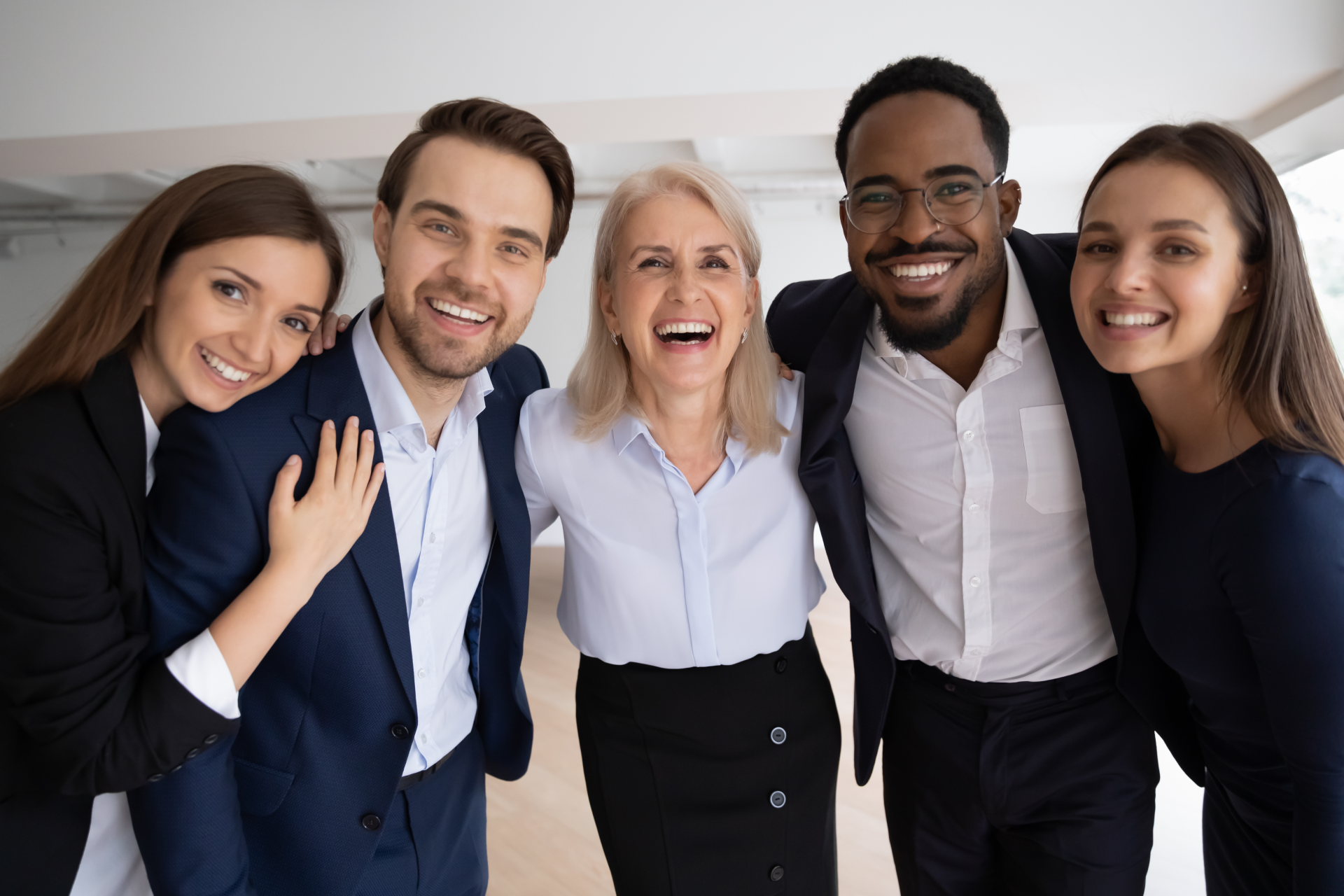 Group of five diverse people smiling and embracing indoors, wearing business attire.