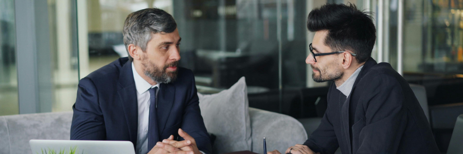 Two men in suits seated, engaged in conversation, in an office setting.