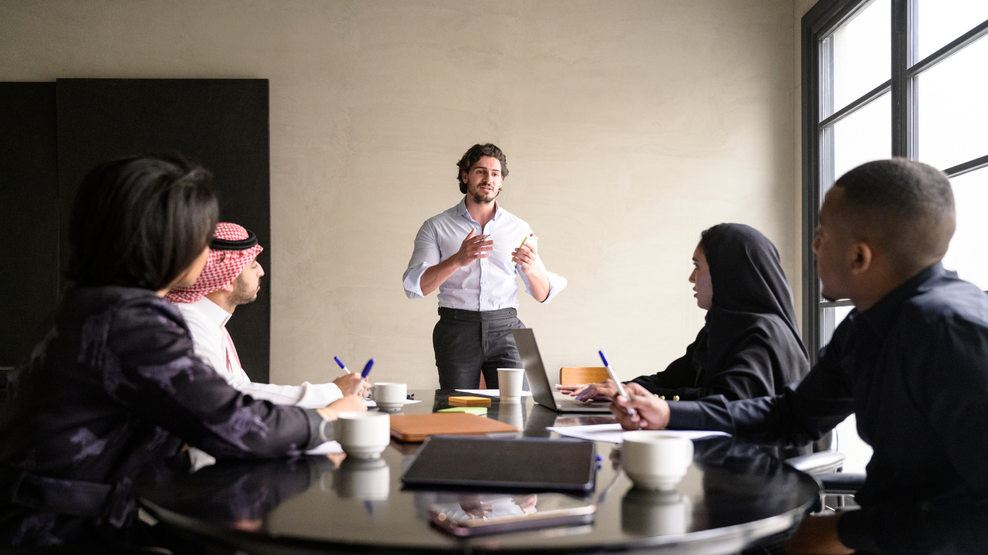 Group in a meeting, one person speaking, gesturing. Others listen, taking notes at a table.