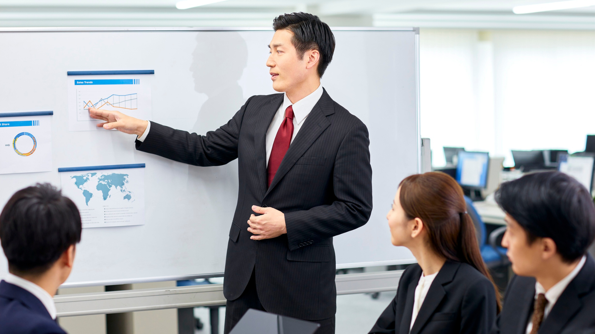 Man in a suit points at a chart on a whiteboard during a presentation to a small group in an office.