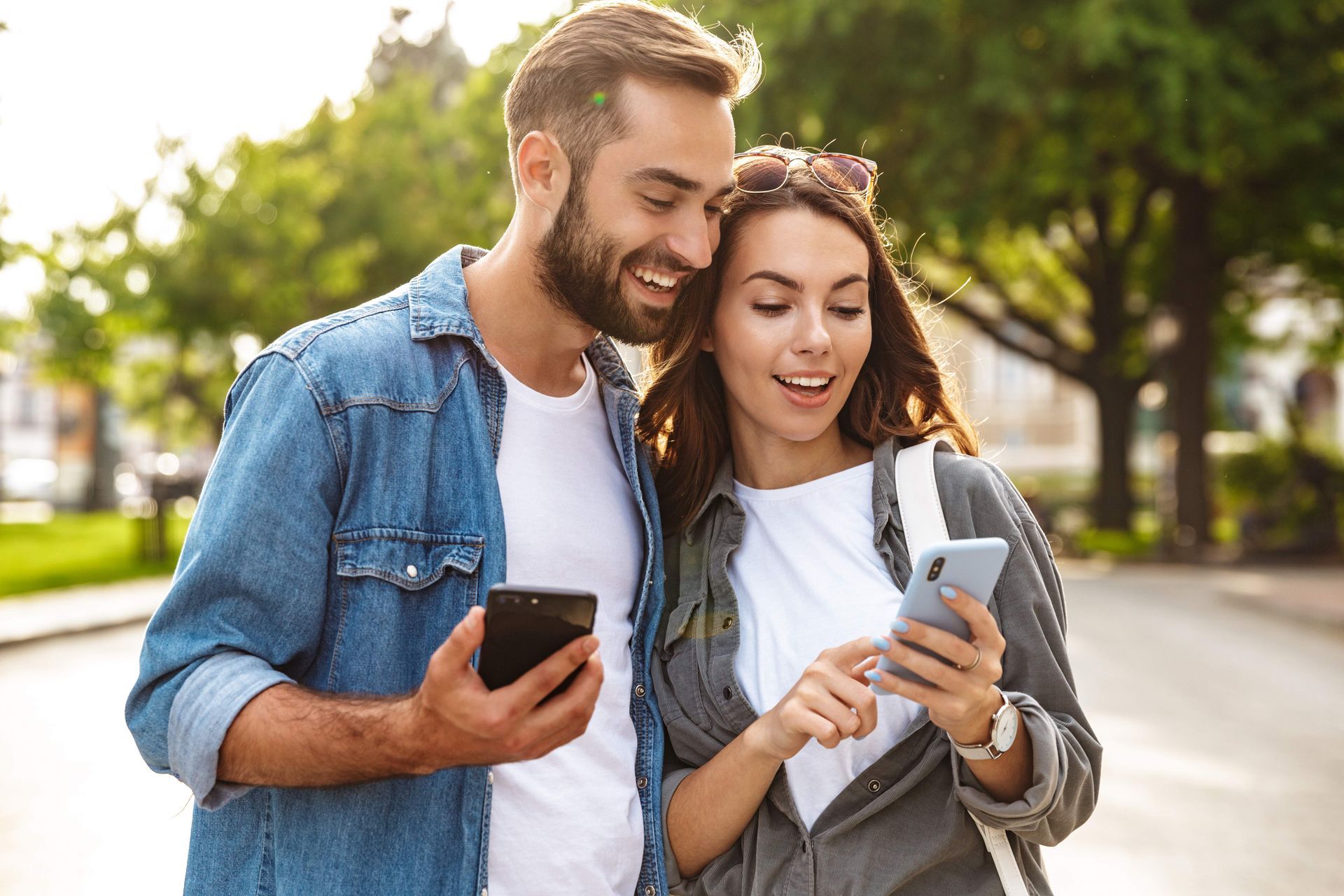Couple looking at phones outdoors; man with beard and denim shirt smiles, woman points at screen.