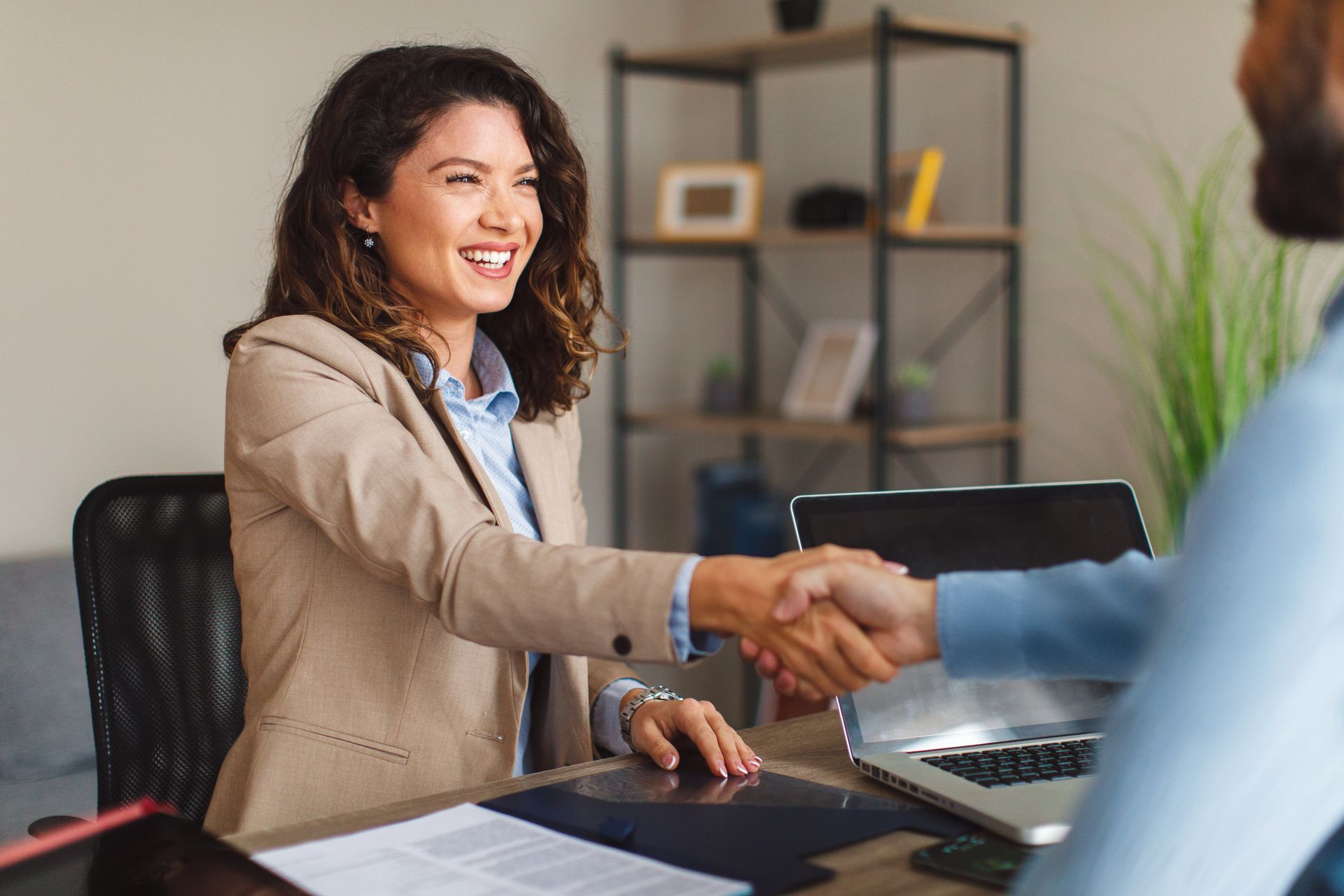 Woman in beige blazer shakes hands with a man at a desk, smiling. Office setting.