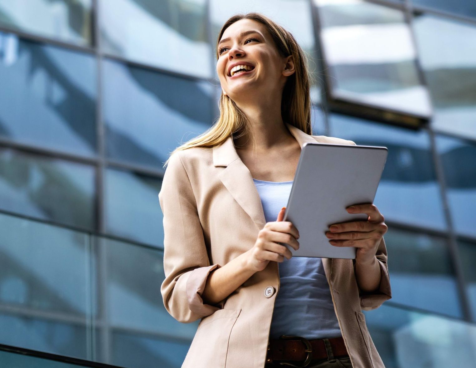 Woman in a tan blazer holding a tablet, smiling, in front of a modern glass building.