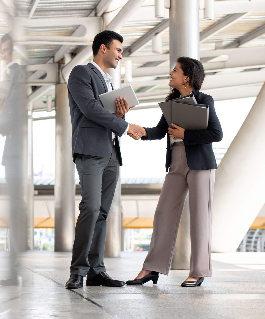 Man and woman in business attire shaking hands, smiling, outdoors in a modern building.
