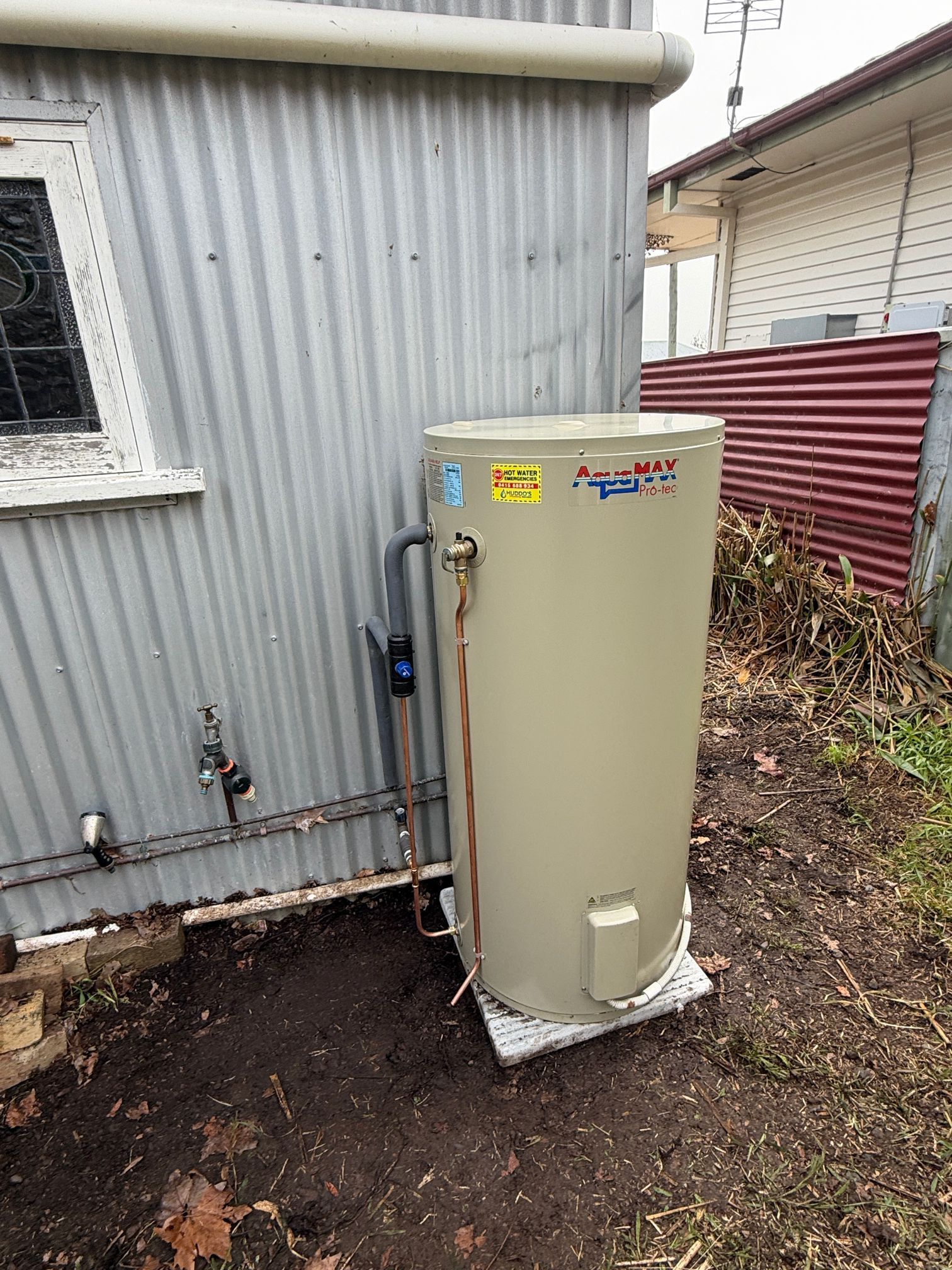 Beige water heater tank beside a grey building, on a concrete pad in a yard. — Huddo's Plumbing Service in Wallalong, NSW