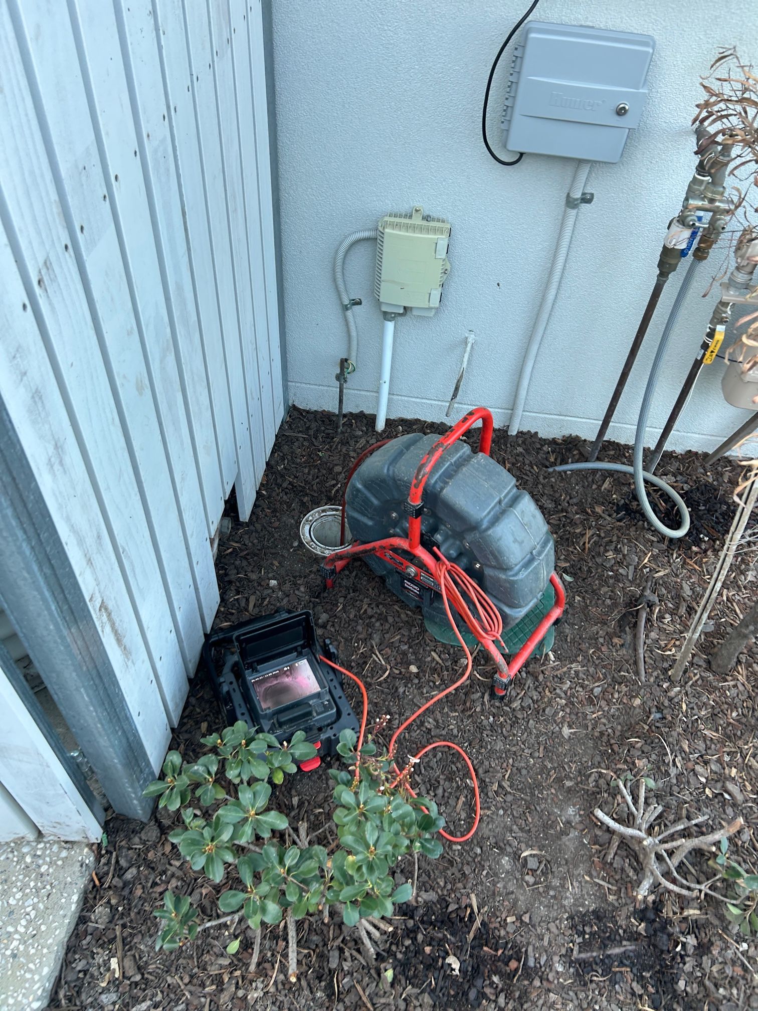 Outdoor utility area with coiled red hoses, a hose reel, and wall-mounted boxes beside a corrugated metal wall — Huddo's Plumbing Service in Wallalong, NSW