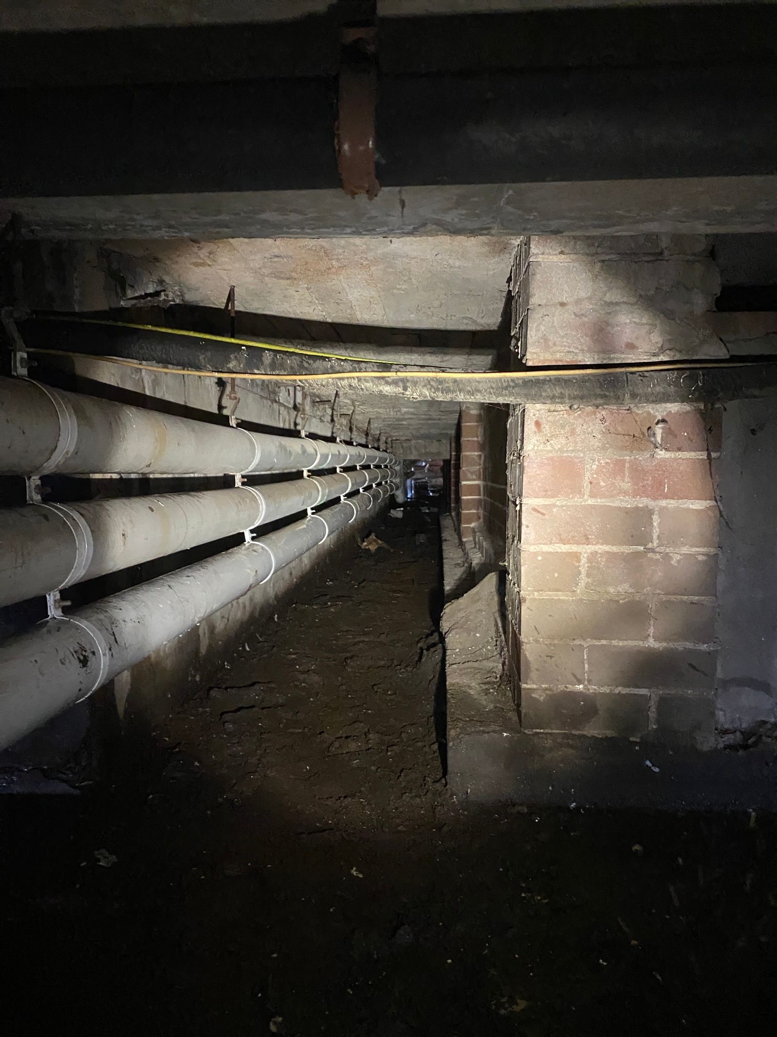 Dim Crawlspace With Pipes Along the Left Wall, a Brick Support Post — Huddo's Plumbing Service in Wallalong, NSW