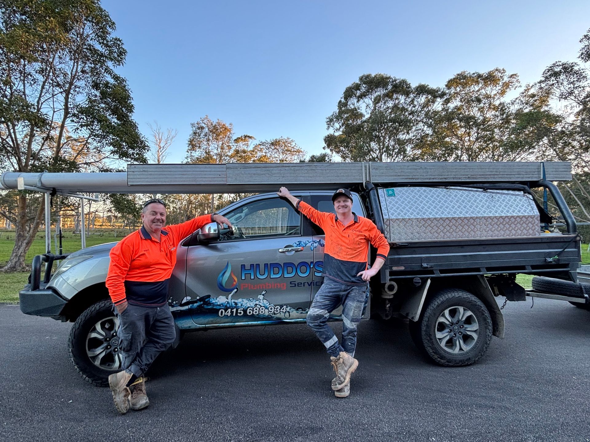 Two People in Orange Shirts Pose — Huddo's Plumbing Service in Wallalong, NSW