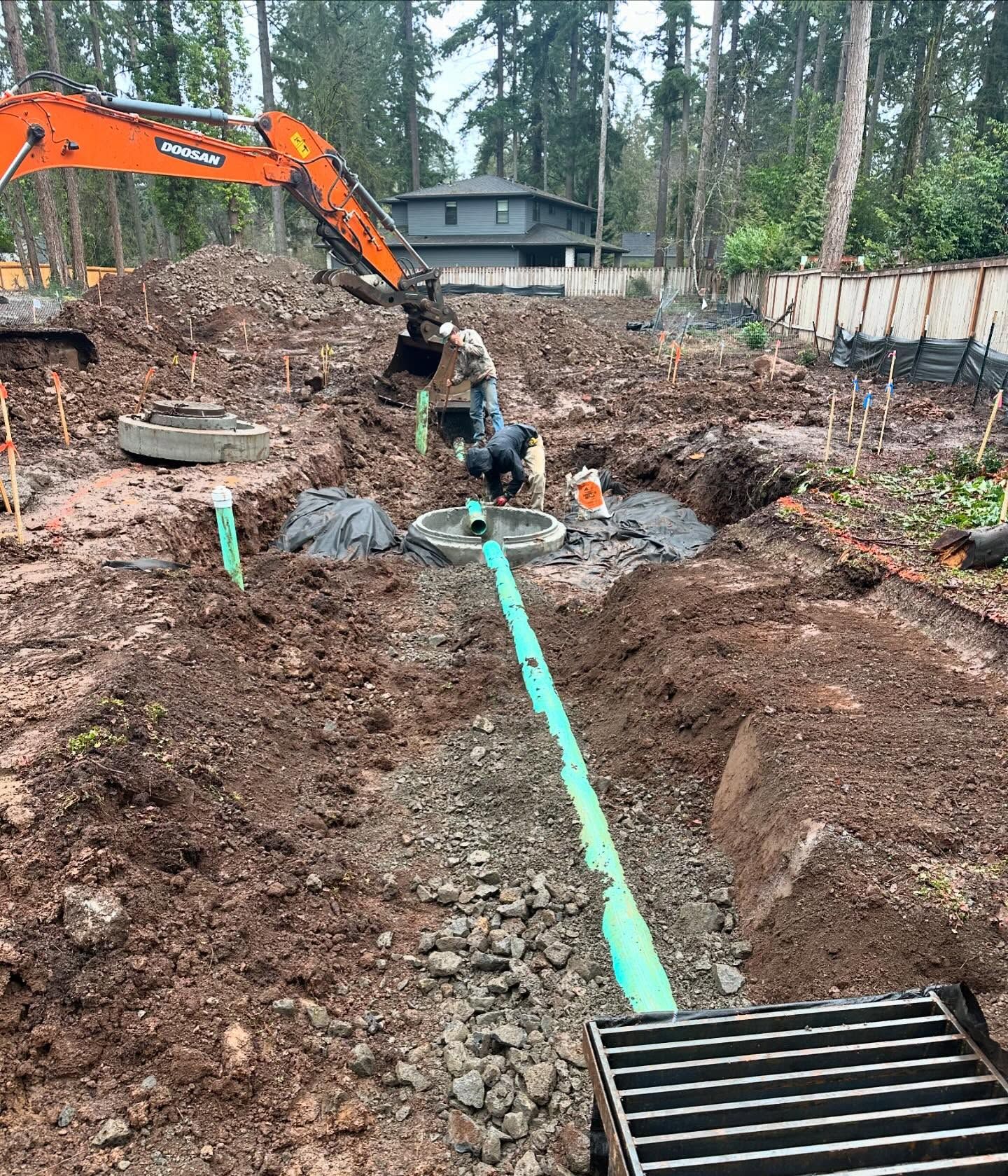 Excavator trenching in a muddy yard beside a house, with green utility lines laid in the ground.