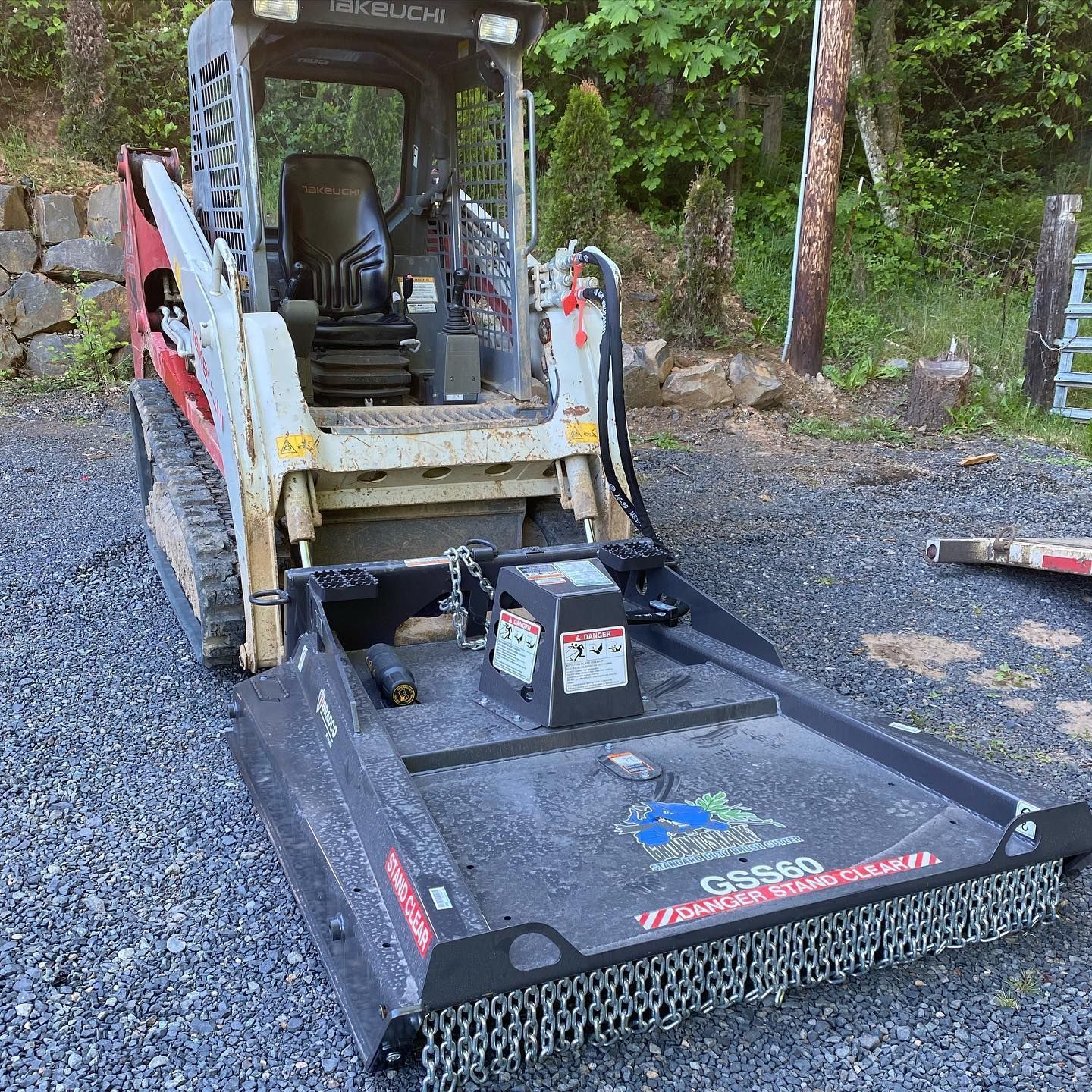 Yellow skid-steer with black forestry mulcher attachment on a gravel driveway by trees.