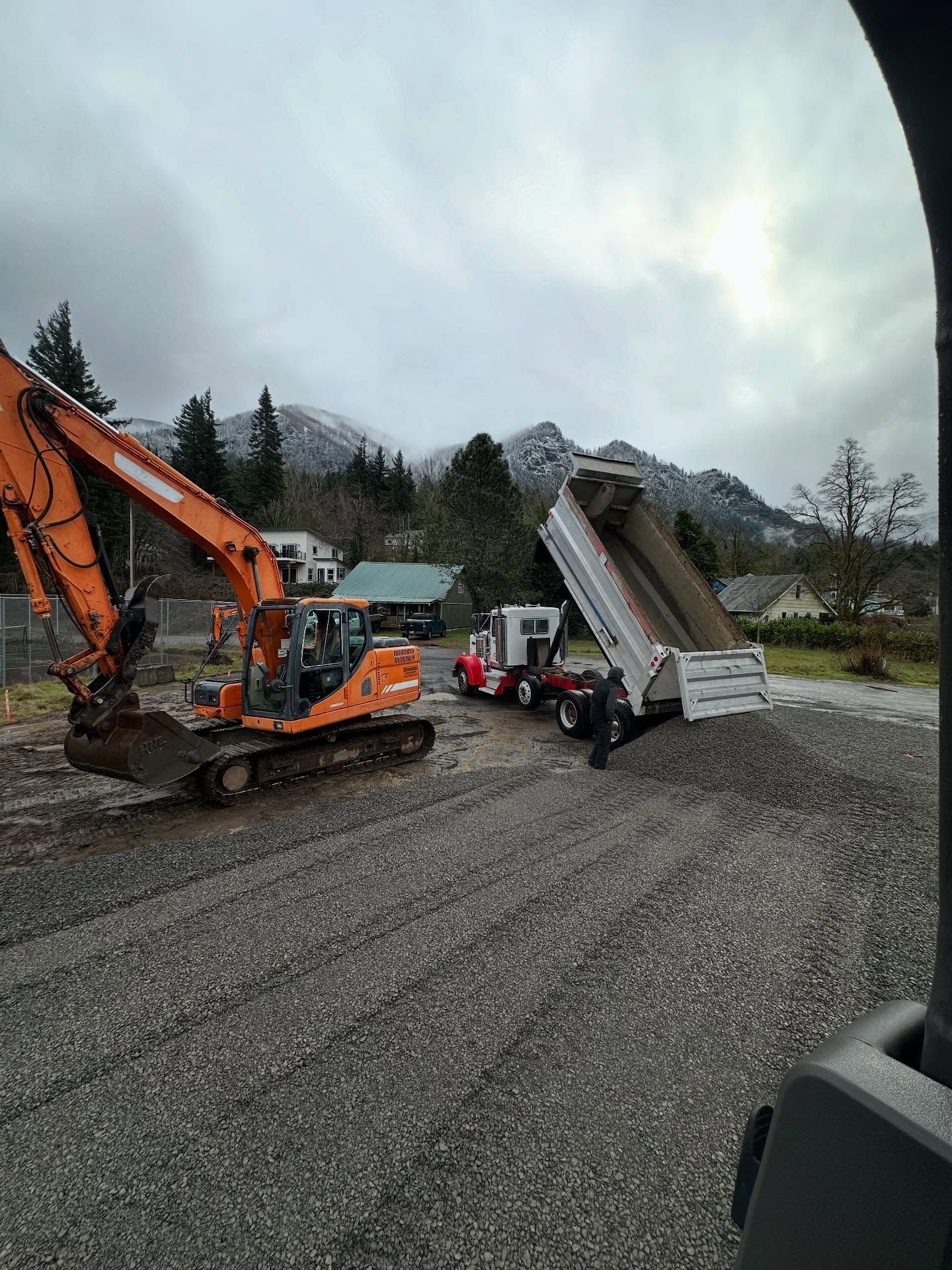 Orange excavator and dump truck tipping gravel on a muddy mountain road under cloudy skies