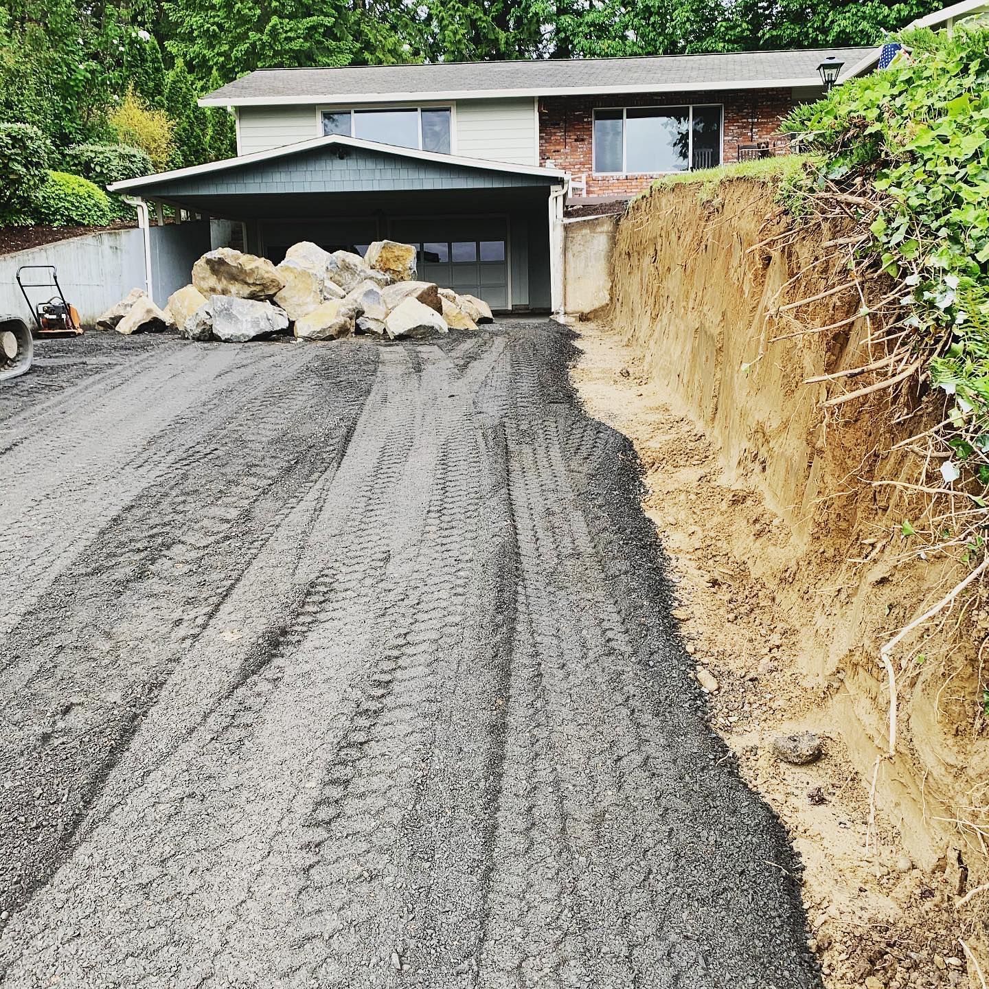 Freshly graded gravel driveway beside a dirt embankment, blocked by large rocks near a house garage.