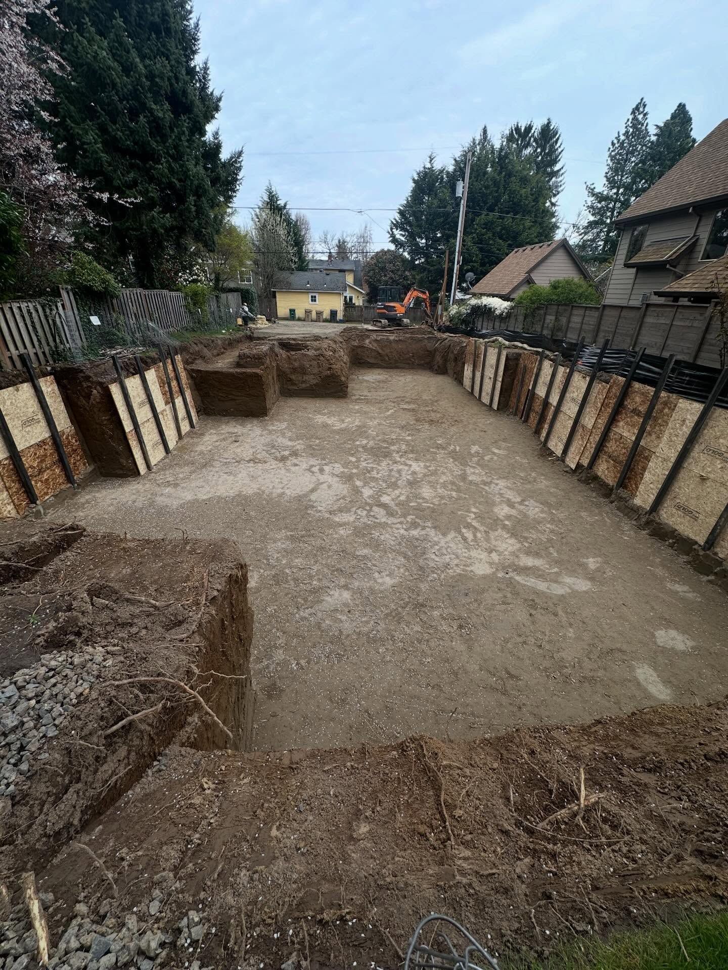 Excavated backyard foundation trench with dirt floor, timber retaining walls, and houses and trees in the background