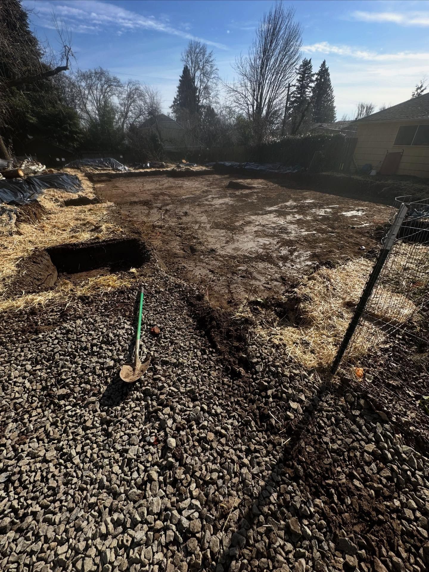 Backyard under construction with gravel, bare ground, a trench, and shovels in bright sunlight