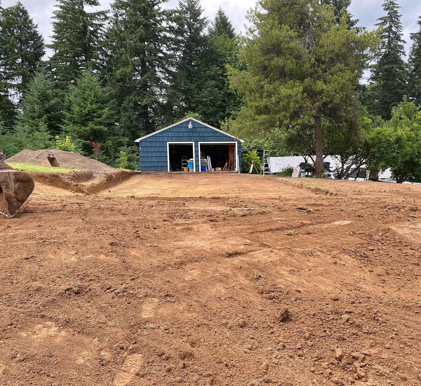 Blue shed at the top of a freshly graded dirt slope, with tall evergreen trees in the background.