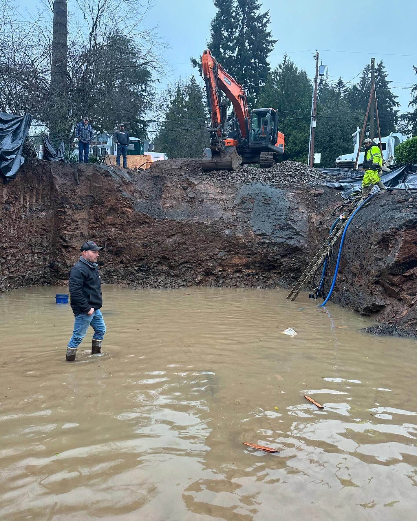 Flooded excavation site with muddy water, an excavator on the bank, and a worker standing in the water