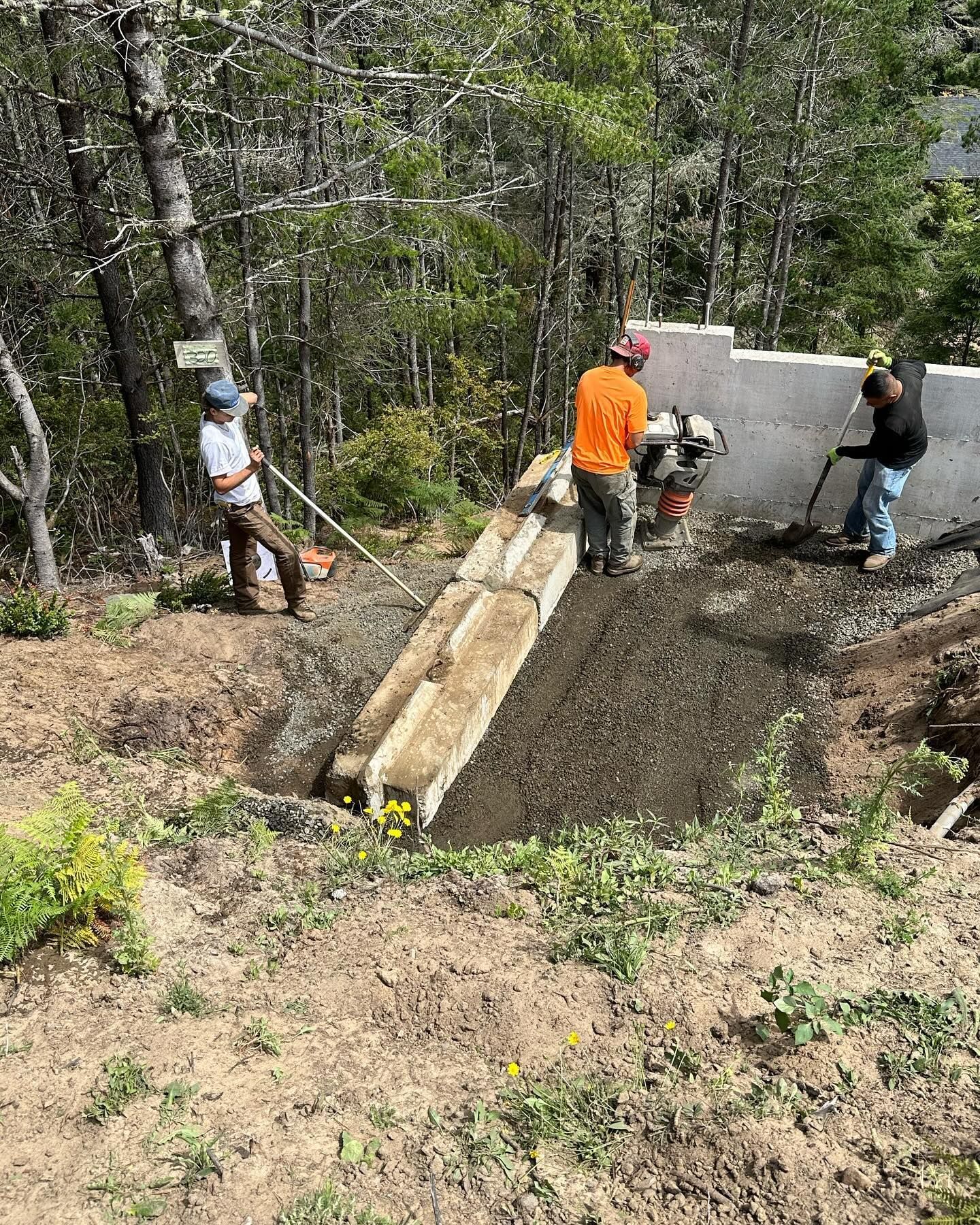 Workers pouring concrete into a foundation in a wooded area, with one guiding the mix from a chute.