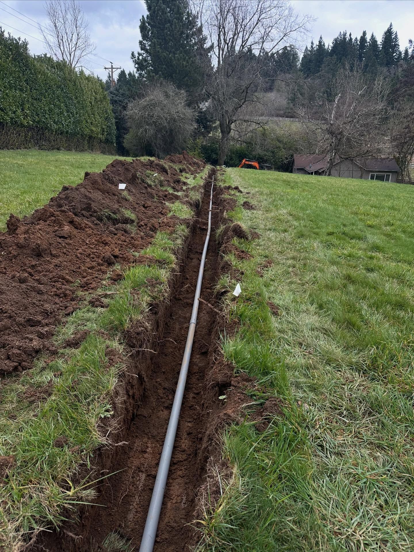 Trenched grassy field with a long black pipe laid in the center, surrounded by soil piles and trees in the background