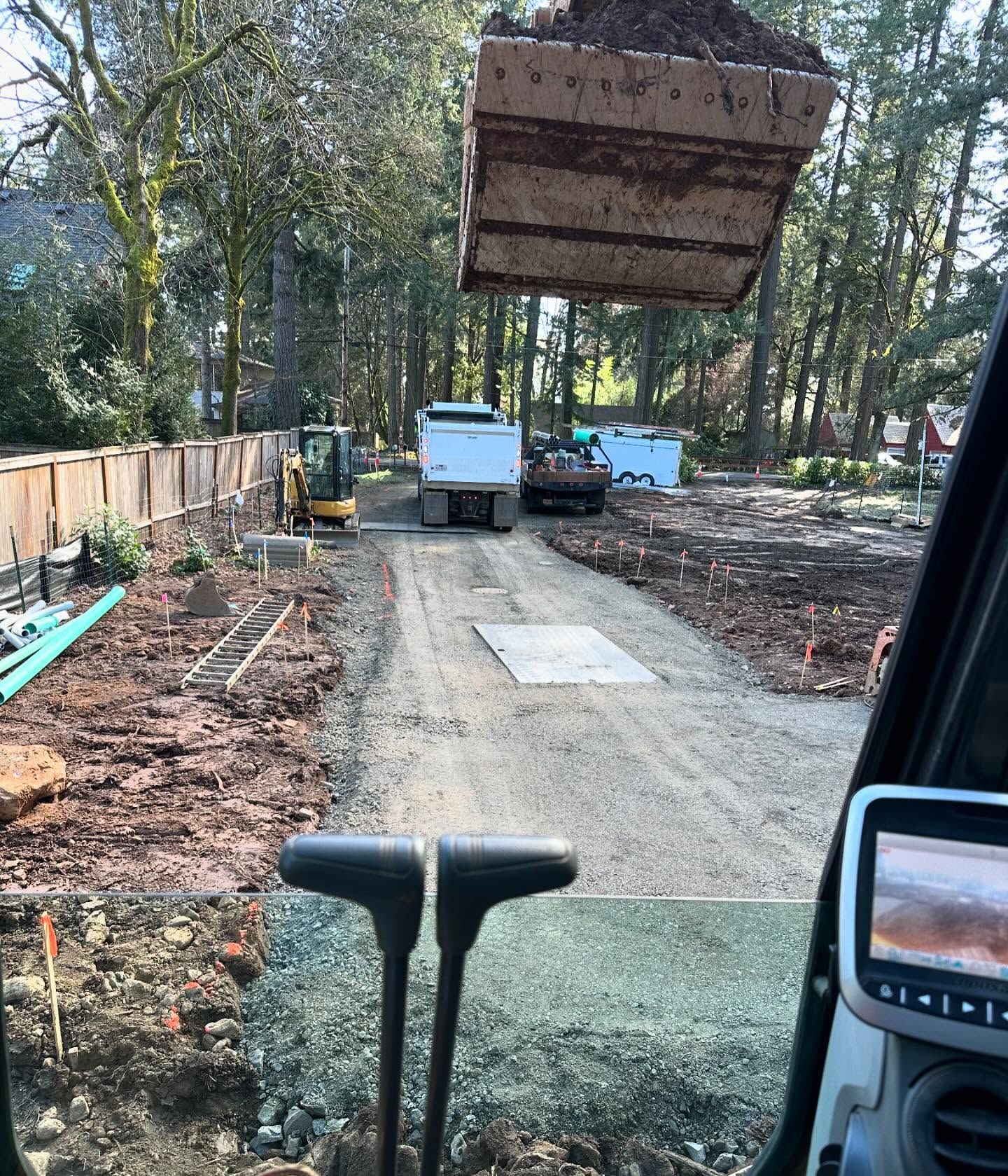 Construction trucks on a dirt driveway under a raised excavator bucket, with trees and fencing nearby.