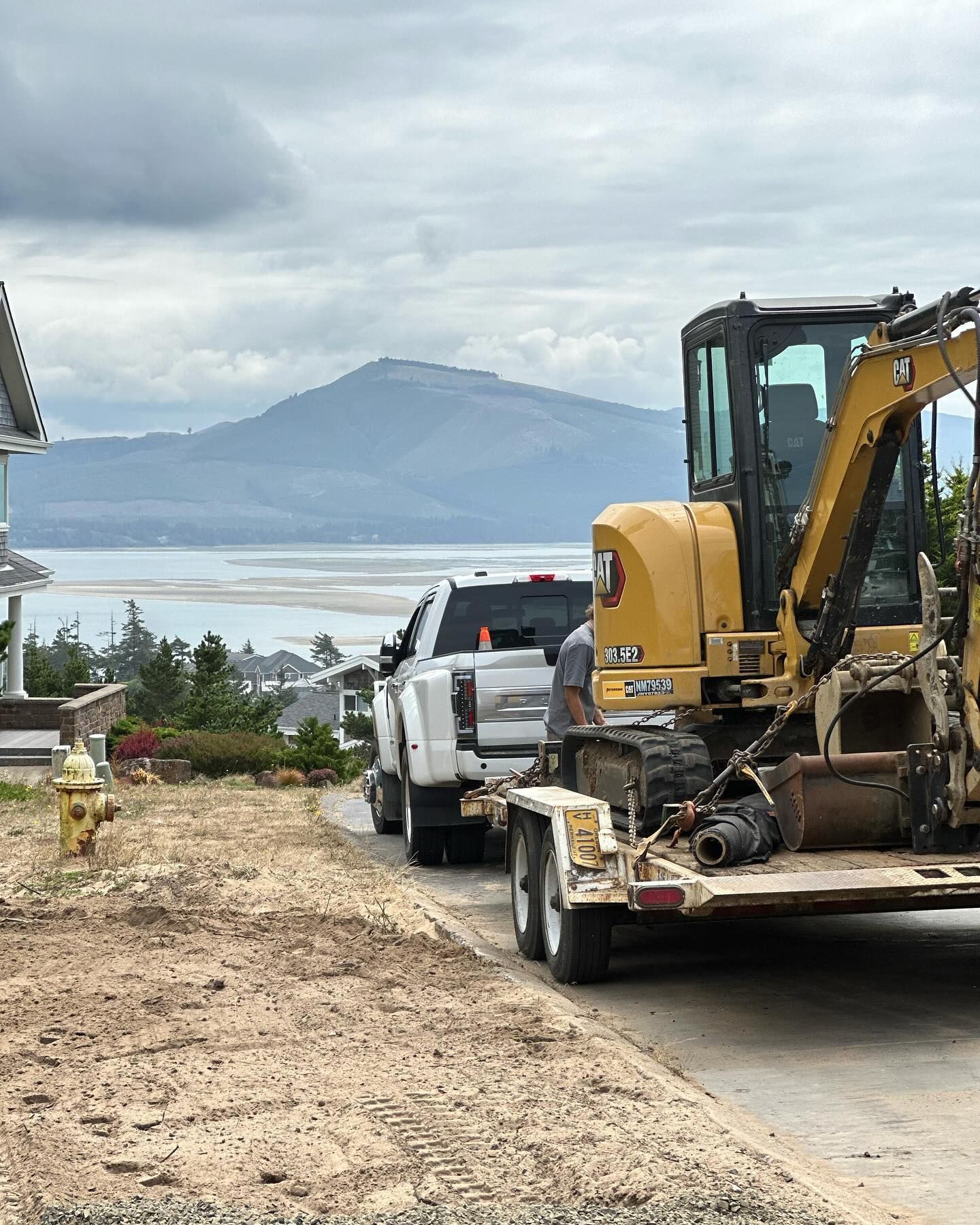 Yellow excavator on a trailer beside a white pickup, overlooking a cloudy mountain and water view