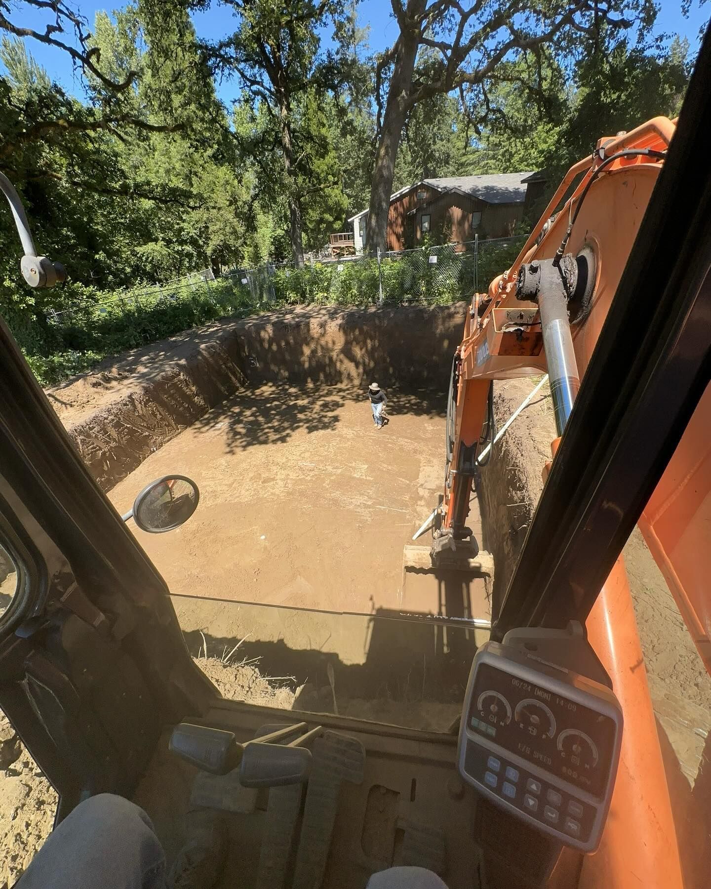 View from a machine cab over muddy water, with an orange excavator arm digging near trees.