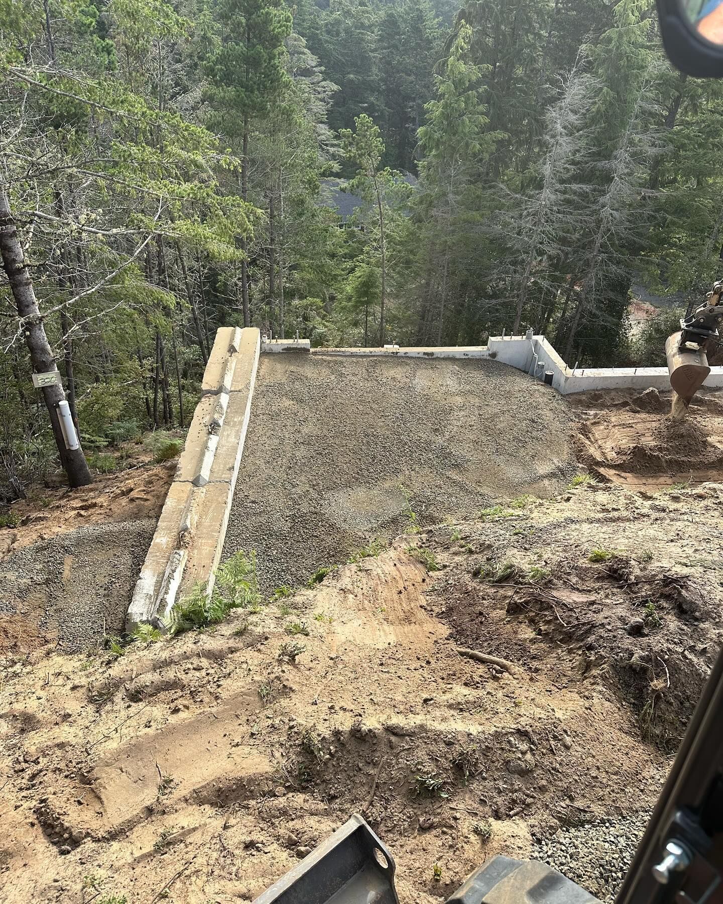 Concrete patio under construction beside a wooded hillside, with gravel and dirt clearing.