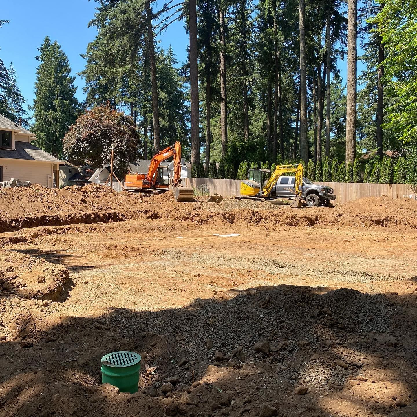 Construction site with two excavators clearing dirt near tall pine trees and a house.