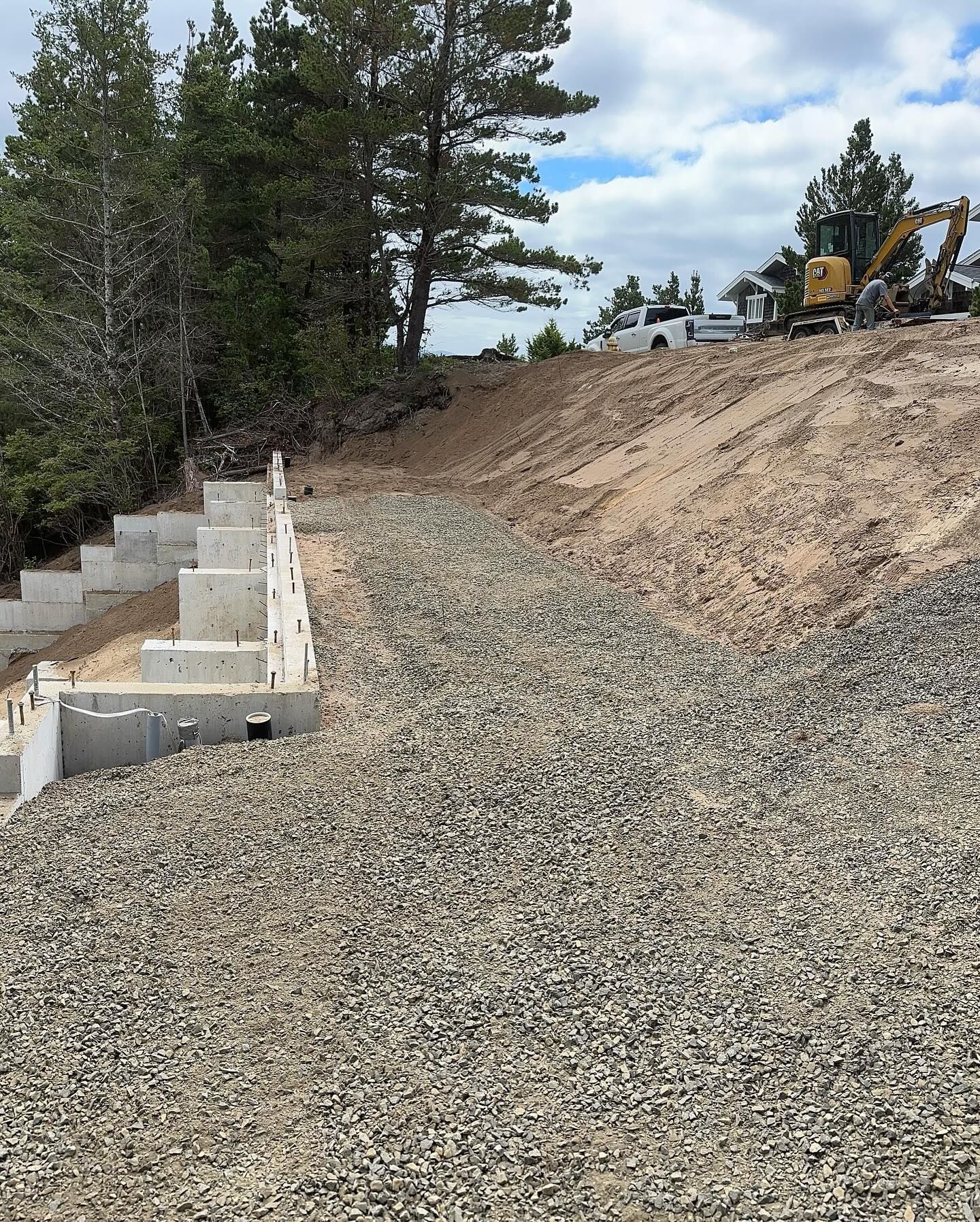 Gravel slope beside concrete retaining steps, with construction equipment on a dirt embankment and trees nearby
